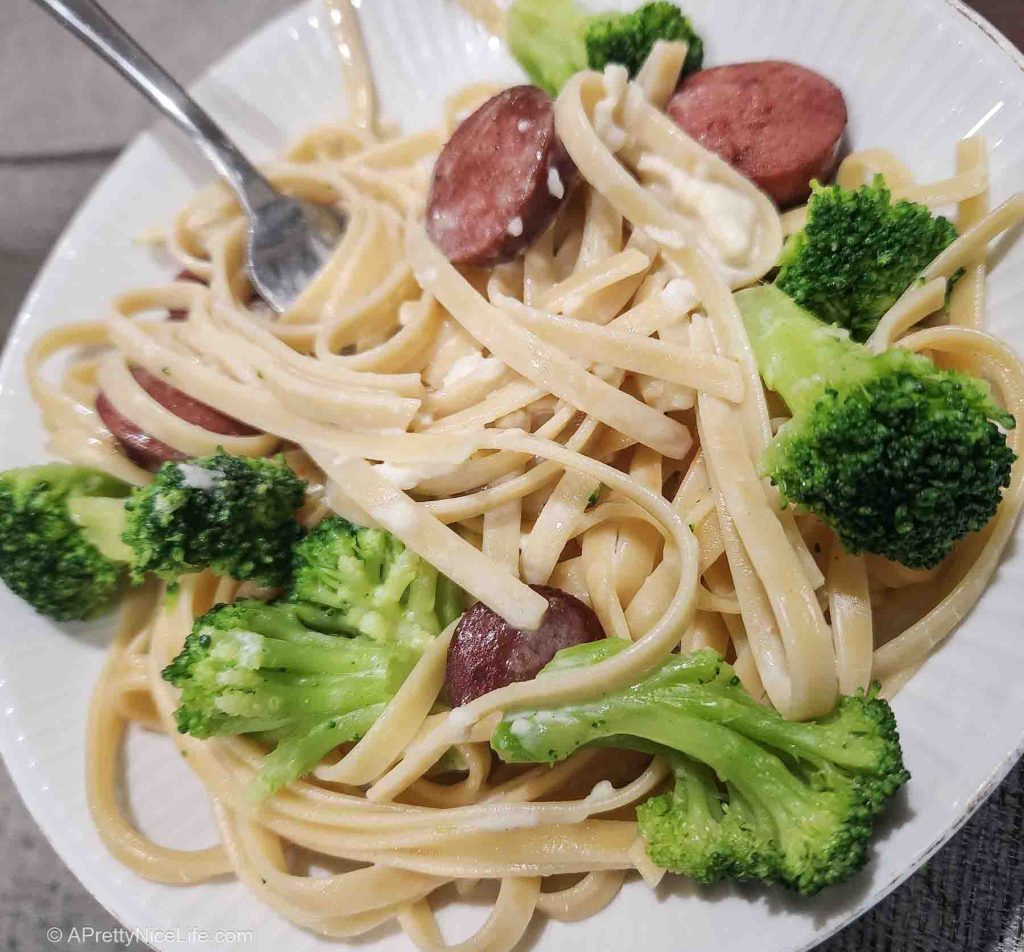 A white china bowl filled with fettucine, alfredo sauce, smoked sausage coins, and bright green broccoli