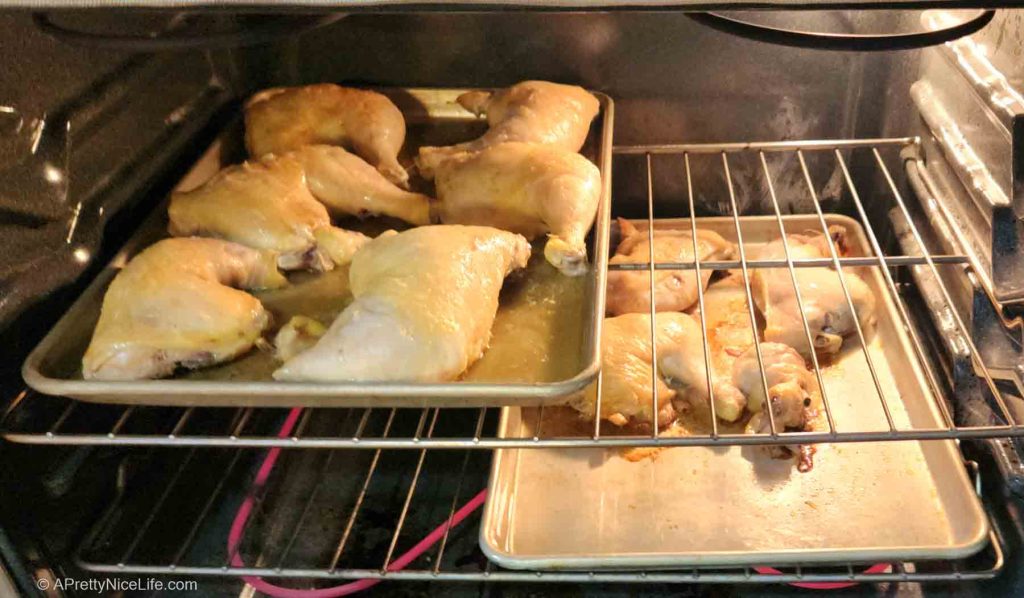 Two aluminum sheet pans with chicken leg quarters roasting in the oven on two racks. 