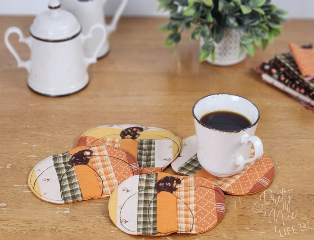 Table set with four cute pumpkin coasters, a cup of coffee, creamer and sugar set, small plant, and some scrap fabric.