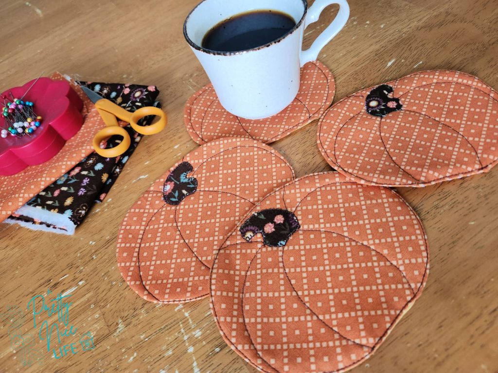 Quilted fabric pumpkin coasters on a wood table with a coffee cup, orange fall fabric scraps, scissors, and a pin cushion