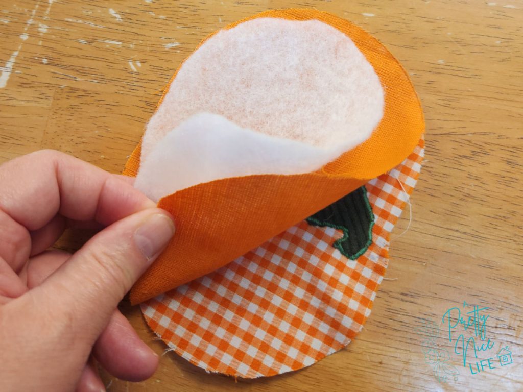 Layers of a quilted pumpkin coaster peeled back to show the gingham front fabric, orange backing, and polyester batting.