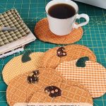 Six quilted fabric pumpkin coasters on a green self-healing cutting mat with a coffee cup, fall fabric scraps, and a sewing machine visible in the corner. Text reads: "Fabric Pumpkin Coasters Sewing Pattern. Make This Cute and Useful DIY Home Decor for Fall"