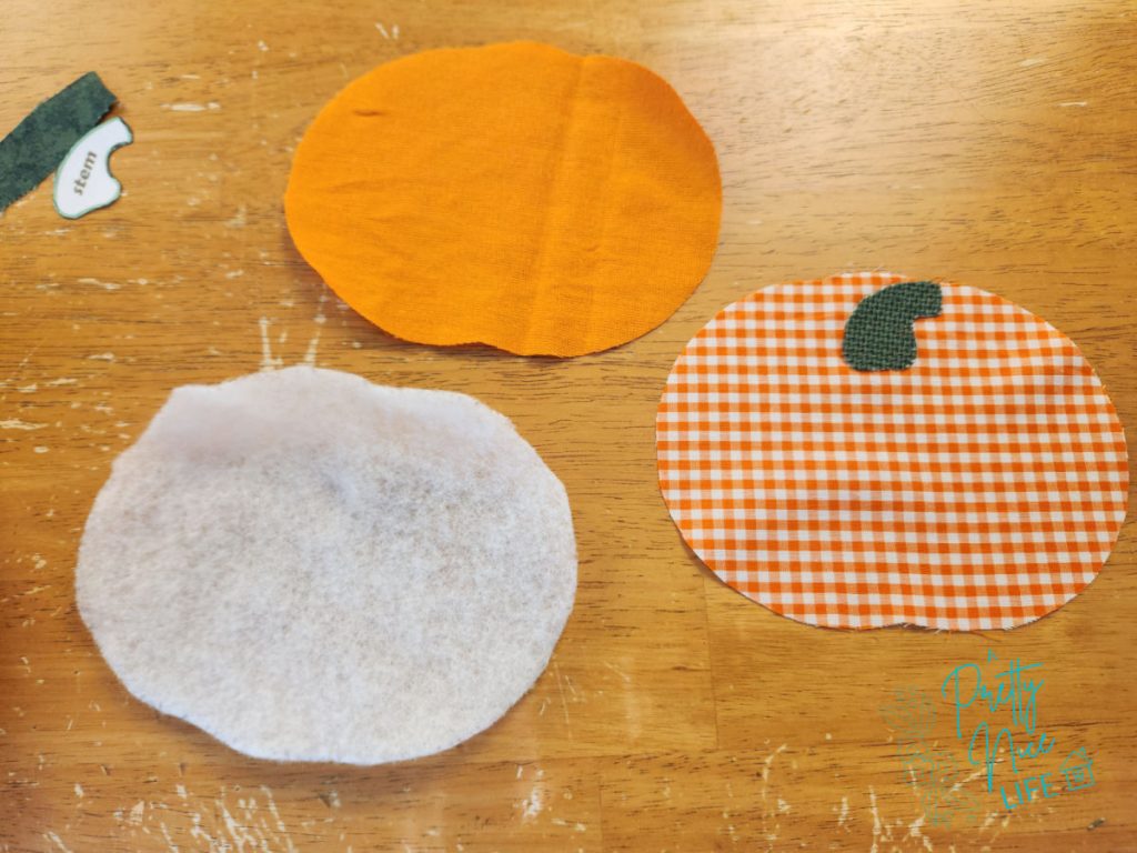 Three cut pieces for a fabric pumpkin coaster — white batting, orange backing, and gingham front with green stem appliquéd on, with paper stem pattern piece nearby