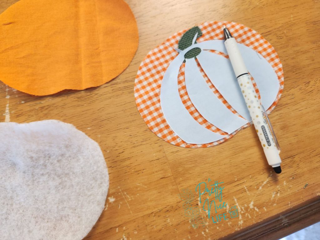Tracing pumpkin rib lines onto a gingham fabric coaster using the pattern piece that is a stencil of where rib lines go and a Frixion heat-erasable pen