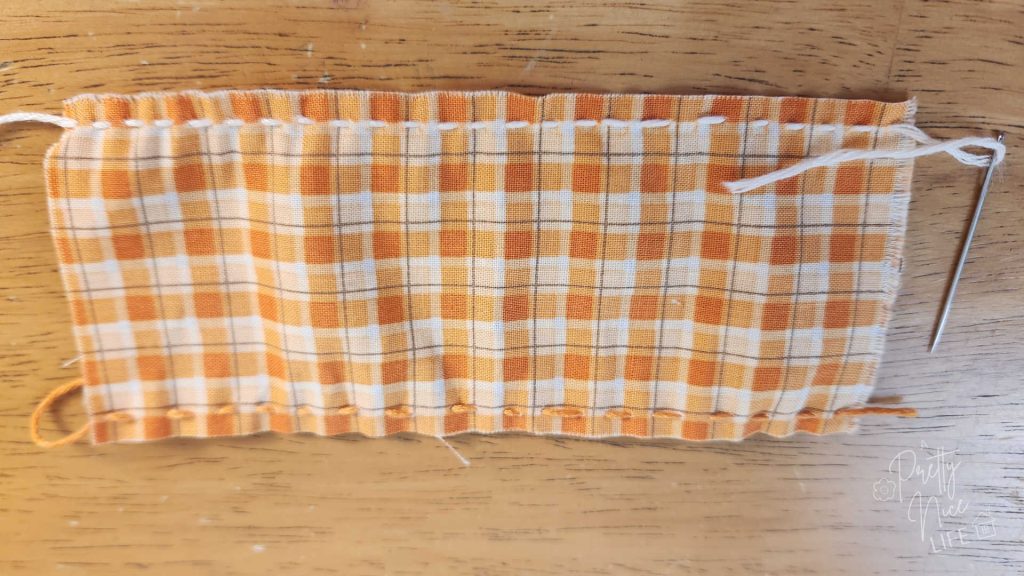Orange and cream colored gingham fabric rectangle laying on a wood table. top and bottom long sides have running stitch. the needle is still attached to the thread at the top.