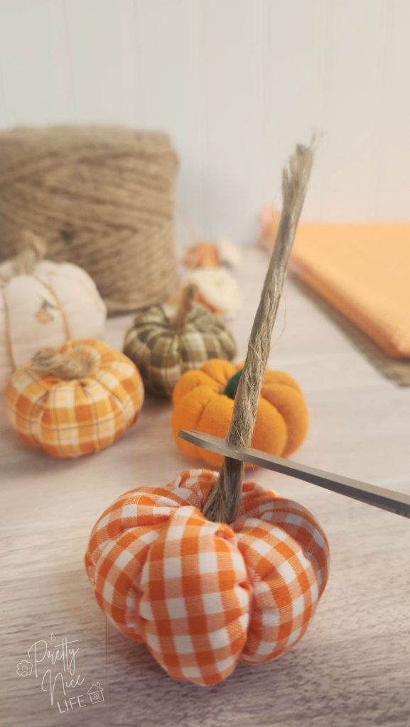 Image shows a small fabric pumpkin with a long stem. Scissors are positioned low on the stem to cut it to the shorter length needed for the fabric pumpkin project.