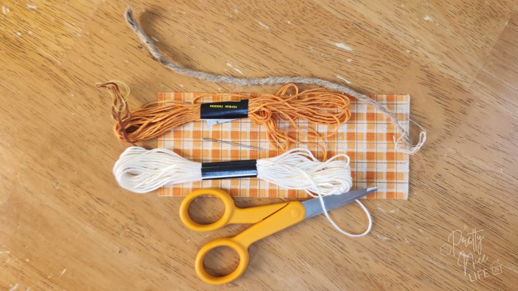 Showing the most basic materials you'll need for making fabric plushie pumpkins: a rectangle of orange and cream colored gingham fabric, embroidery floss, a tapestry needle, scissors, and a small length of twine. 