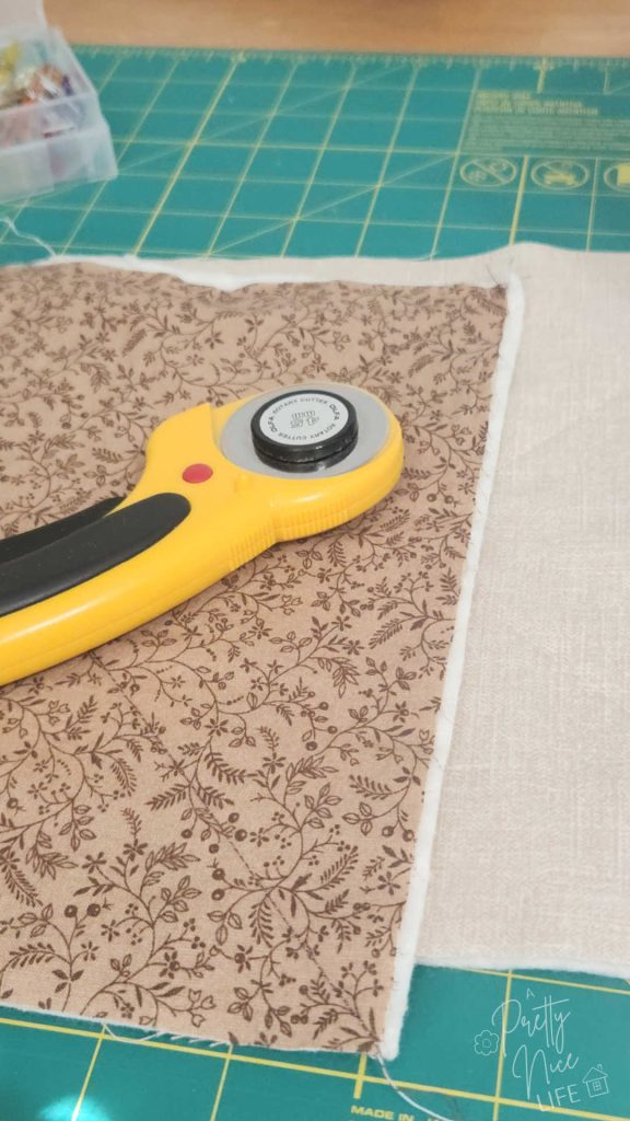 Shows closeup of rotary cutter laying on cotton fabric, 100% cotton batting, and cotton thread for sewing a microwavable bowl cozy on top of green self-healing cutting mat, with a box of sewing clips in the background
